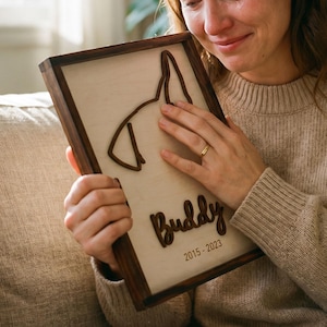 Puede incluir: Placa conmemorativa de madera con silueta de perro. La placa incluye el nombre "Buddy" y los años "2015-2023". El marco es marrón oscuro y la silueta del perro es de un tono de madera más claro.