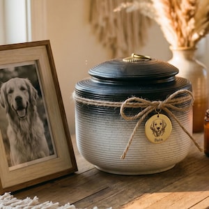 May include: A wooden framed photo of a Golden Retriever next to a black and white pet urn with a gold pendant. A lit candle sits nearby on a wooden table. The urn has a rope bow and a gold pendant with a dog illustration.