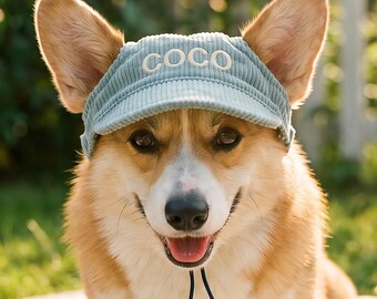 May include: A light blue corduroy visor with the word COCO embroidered in white thread on the front. The visor features a dark blue adjustable chin strap and is worn by a tan and white Corgi dog with large upright ears and a happy expression.