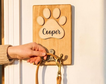May include: A rectangular wooden wall-mounted leash holder featuring a carved paw print design and the name Cooper engraved in black script. A brass-colored metal hook is attached to the center, holding a tan leather leash against a white wall.