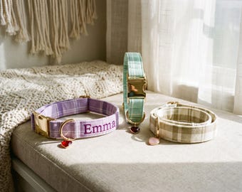 May include: Three fabric pet collars with gold-tone metal buckles and D-rings. One lavender plaid collar features the name Emma embroidered in purple. One mint green plaid collar stands upright. One beige plaid collar is coiled with a heart charm.