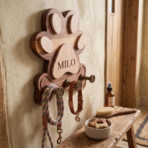 Puede incluir: Un soporte de pared de madera para correas de mascotas con forma de huella y el nombre MILO grabado en la almohadilla central. La placa de madera oscura cuenta con tres ganchos de metal color bronce, cada uno con un dise&ntilde;o de huella peque&ntilde;a.