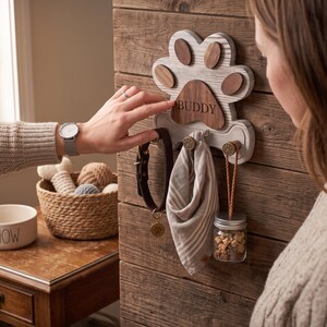 Puede incluir: Un soporte de pared para correas de mascotas hecho de madera con forma de huella y acabado blanco desgastado. La almohadilla central es de madera oscura grabada con el nombre BUDDY. Incluye tres ganchos met&aacute;licos con un collar marr&oacute;n, pa&ntilde;uelo y frasco.