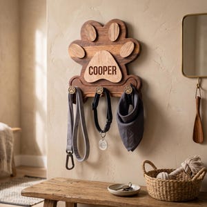 Puede incluir: Un soporte de pared para correas de mascotas hecho de madera con forma de huella. La base de madera oscura presenta un centro de madera clara grabado con el nombre COOPER. Tres peque&ntilde;os ganchos con detalles de huellas sostienen una correa gris.