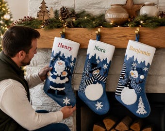 May include: Three blue Christmas stockings with white cuffs, each featuring a unique embroidered character: a Santa Claus, a snowman with a striped scarf, and a snowman with a blue patterned scarf. The names Henry, Nora, and Benjamin are embroidered.