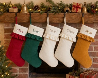 May include: Five velvet Christmas stockings with cream-colored cuffs and ruffled trim. The stockings are red, green, cream, cream, and mustard yellow. Each cuff is embroidered with a name: Alice, Liam, Isaac, Claire, and Mom.