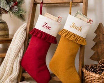 May include: Two velvet Christmas stockings hanging from a wooden ladder. The left stocking is deep red with a white cuff embroidered with the name Zoe. The right stocking is mustard yellow with a white cuff embroidered with the name Jack.