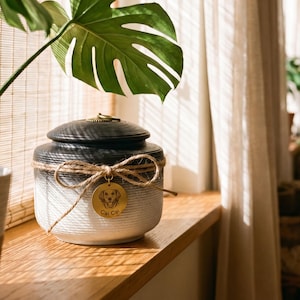 May include: A ceramic pet urn with a black lid and a white and gray body. The urn is adorned with a twine bow and a gold tag with a dog illustration and the name "Cal Cal". The urn is sitting on a wooden surface.