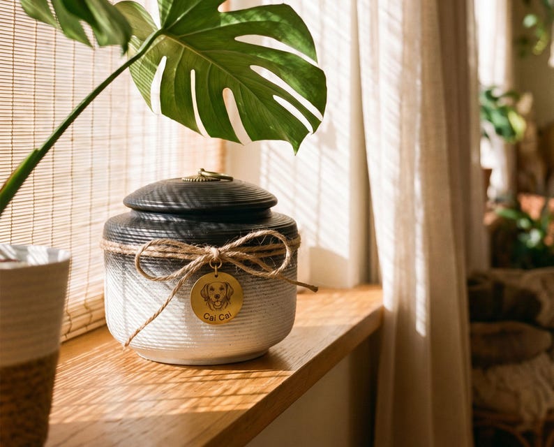 May include: A ceramic pet urn with a black lid and a white and gray body. The urn is adorned with a twine bow and a gold tag with a dog illustration and the name "Cal Cal". The urn is sitting on a wooden surface.