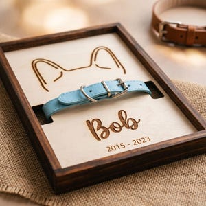 Puede incluir: Una caja de sombra de madera con un collar para perro azul claro. La caja tiene un interior de madera clara con un diseño de oreja de perro tallado y el nombre "Bob" con los años "2015-2023". Un collar de perro de cuero marrón está en el fondo.