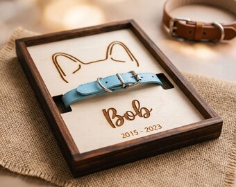 May include: A wooden shadow box with a light blue dog collar. The box has a light wood interior with a carved dog ear design and the name "Bob" with the years "2015-2023". A brown leather dog collar is in the background.