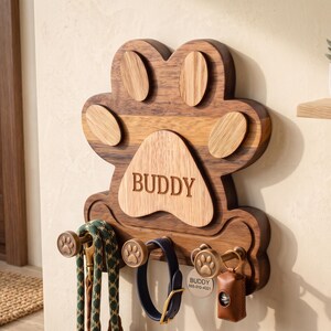 Puede incluir: Un soporte de pared para correas de mascotas hecho de madera con forma de huella. La base de madera oscura tiene un centro de madera m&aacute;s clara grabado con el nombre BUDDY. Incluye tres ganchos color bronce con detalles de huellas para colgar.
