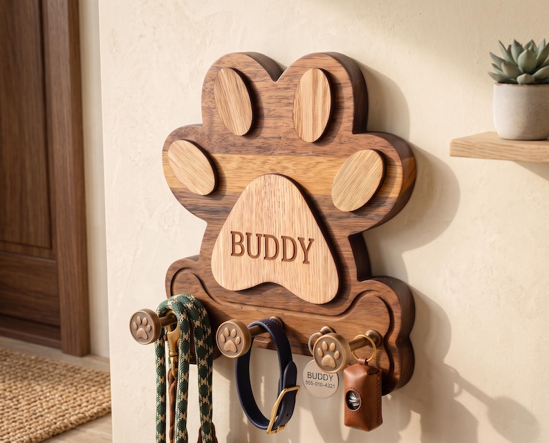Puede incluir: Un soporte de pared para correas de mascotas hecho de madera con forma de huella. La base de madera oscura tiene un centro de madera m&aacute;s clara grabado con el nombre BUDDY. Incluye tres ganchos color bronce con detalles de huellas para colgar.