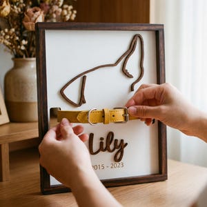 Puede incluir: Un marco conmemorativo de madera para mascotas con una silueta de perro, el nombre "Lily" y fechas. Se está colocando un collar de perro de cuero amarillo en el marco. El marco es marrón con un fondo blanco.