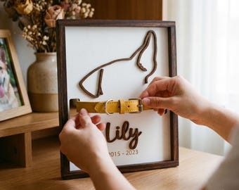 May include: A framed wooden pet memorial with a dog silhouette and the name "Lily" and dates. A yellow leather dog collar is being placed in the frame. The frame is brown with a white background.