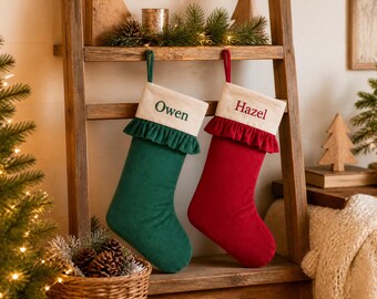 May include: Two Christmas stockings hanging side by side. The left stocking is dark green with a cream cuff featuring the name Owen in green embroidery and a green ruffle. The right stocking is red with a cream cuff featuring the name Hazel in red embroidery and a red ruffle.
