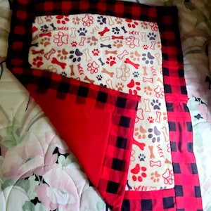 May include: A dog bed with a red and black buffalo plaid border. The center features a cream-colored fabric with paw prints, bones, and the word "Woof" in red, brown, and blue. The bed is folded, revealing a solid red underside.