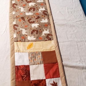May include: A rectangular quilted table runner featuring a patchwork design with autumn-themed fabrics in shades of brown, orange, red, and white. The center panel displays white silhouettes of bears and deer among brown leaves on a tan background.