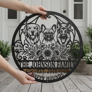 Könnte beinhalten: Ein rundes Wandbild aus schwarzem Metall mit lasergeschnittenen Porträts von drei Hunden, darunter ein Golden Retriever, eine Französische Bulldogge und ein Deutscher Schäferhund, umgeben von Blumenmotiven und Pfotenabdrücken, mit dem Text THE JOHNSON FAMILY EST. 2018 HAPPY HOUNDS.