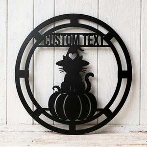 Könnte beinhalten: Ein rundes Wandbild aus schwarzem Metall mit der Silhouette einer Katze, die einen Hexenhut mit herzförmigem Ausschnitt trägt und auf einem Kürbis sitzt. Im oberen Teil des kreisförmigen Rahmens befindet sich der Text CUSTOM TEXT.