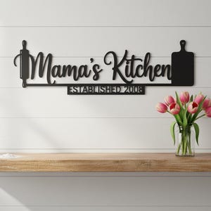 Könnte beinhalten: Schwarzes Küchenschild aus Metall mit den Worten "Mama's Kitchen" in Schreibschrift, einer Teigrolle und einer Schneidebrett-Silhouette. Das Schild enthält auch den Text "ESTABLISHED 2008".