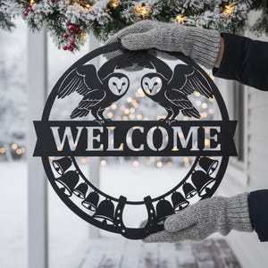 Könnte beinhalten: Ein kreisförmiges Wand-Schild aus schwarzem Metall mit zwei Schleiereulen im oberen Bereich, einem mittigen horizontalen Banner mit dem ausgeschnittenen Wort WELCOME sowie einer dekorativen Bordüre aus Glocken und einem Hufeisen.