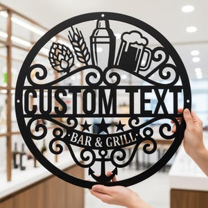 Puede incluir: Letrero de bar de metal negro con un diseño circular. El letrero presenta las palabras "CUSTOM TEXT" y "BAR & GRILL", junto con imágenes de una jarra de cerveza, una coctelera, lúpulo y trigo. También se incluye un ancla y estrellas.