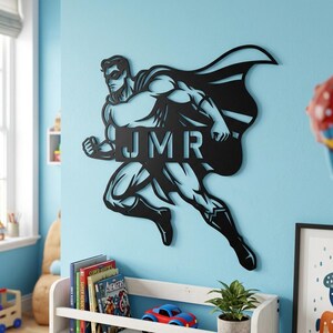 Op de afbeelding: Zwarte metalen wandkunst van een superheld met de initialen "JMR". De superheld is afgebeeld in een rennende pose met een cape. Gemonteerd op een lichtblauwe muur, ideaal voor een kinderkamer. Ongeveer 60 cm hoog.