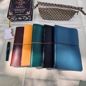 Pode incluir: Cinco diários encadernados em couro nas cores bordô, laranja, verde, preto e azul, cada um preso com um elástico. Ao lado, há uma caneta-tinteiro preta, um bloco de notas em forma de gato, um livro intitulado De La Chair Et Du Sang e um estojo estampado.