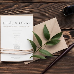 Könnte beinhalten: Hochzeitseinladungs-Set mit einer cremefarbenen Einladung mit den Namen "Emily & Oliver" und dem Datum "14. September 2025, Florenz, Italien". Die Einladung ist mit Bindfaden gebunden. Ein brauner Umschlag, grüne Blätter, ein Stift und Tinte sind ebenfalls vorhanden.