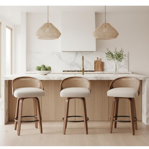 May include: Three wooden bar stools with woven backs and cream-colored seats are arranged in front of a kitchen island. The stools have a curved back design and a footrest. Two woven pendant lights hang above the island. The kitchen has light wood cabinets and a marble countertop.