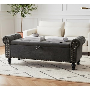May include: A dark gray storage bench with a tufted top and rolled arms. The bench is accented with brass-colored nailhead trim and sits on four black, turned legs. Books and a small bowl rest on top of the bench.