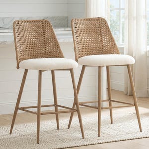 May include: Two woven rattan bar stools with cream-colored, rounded seats. The backs of the stools are woven in a grid pattern. The legs are a light brown color and have a footrest. The stools are placed on a light-colored rug.