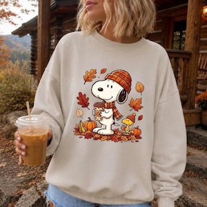 May include: A beige crewneck sweatshirt featuring a graphic print of a white cartoon dog wearing an orange plaid beanie and scarf, holding a coffee cup. The design includes a small yellow bird, orange pumpkins, and scattered autumn leaves.