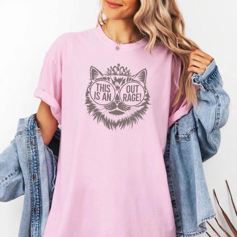 May include: Pink t-shirt featuring a cat graphic wearing sunglasses and a crown. The graphic includes the text "THIS IS AN OUT RAGE!". The shirt has short sleeves and a relaxed fit.