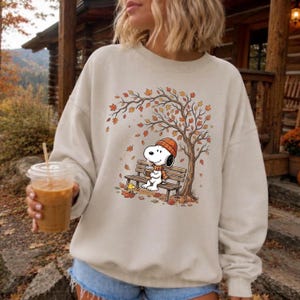 May include: A beige crewneck sweatshirt featuring a graphic print of a white cartoon dog wearing an orange beanie and scarf, sitting on a wooden bench under an autumn tree with falling orange leaves, while holding a cup of coffee.