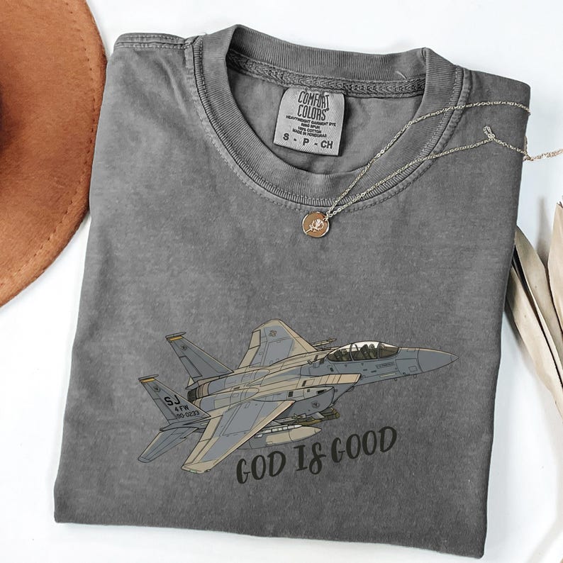 Pu&ograve; includere: Una maglietta grigia Comfort Colors con un'illustrazione di un jet da combattimento e la scritta "GOD IS GOOD". La maglietta ha un girocollo e maniche corte. Una collana d'argento con un ciondolo &egrave; drappeggiata sulla maglietta.