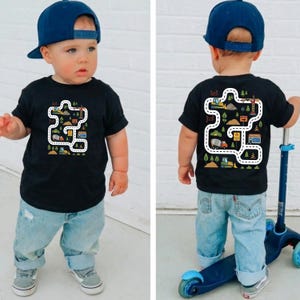 May include: Black toddler t-shirt with a colorful road and construction vehicle design. The design is on the front and back of the shirt. The child is wearing a blue baseball cap, light blue jeans, and gray sneakers.