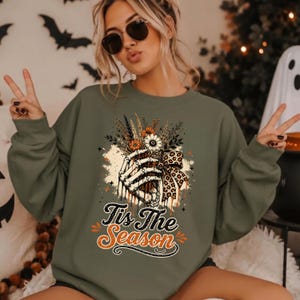 May include: An olive green crewneck sweatshirt featuring a graphic print of a skeletal hand holding a bouquet of flowers with a leopard print bow. The text "Tis The Season" is printed below the graphic in a stylized black and orange font.