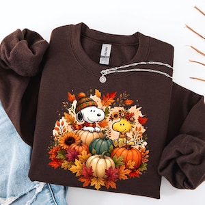 May include: A dark brown crewneck sweatshirt featuring a graphic print of Snoopy wearing a striped beanie and scarf sitting on a pumpkin, alongside Woodstock wearing a floral crown, surrounded by colorful autumn leaves and various pumpkins.