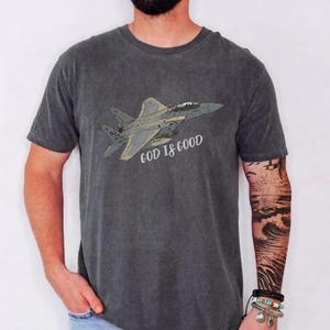 Pu&ograve; includere: T-shirt grigio scuro con un'illustrazione di un jet da combattimento e la scritta "GOD IS GOOD". Il design &egrave; in tonalit&agrave; di grigio e beige. La maglietta &egrave; in materiale morbido.