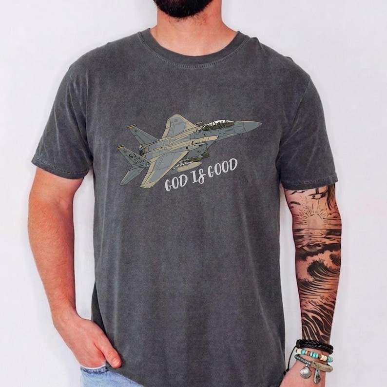 Pu&ograve; includere: T-shirt grigio scuro con un'illustrazione di un jet da combattimento e la scritta "GOD IS GOOD". Il design &egrave; in tonalit&agrave; di grigio e beige. La maglietta &egrave; in materiale morbido.