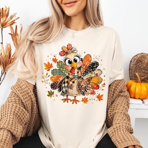 May include: A cream-colored t-shirt featuring a central graphic of a cartoon turkey with large eyes and long eyelashes. The turkey is wearing a plaid scarf and has colorful, patterned feathers in shades of orange, brown, and black, surrounded by scattered autumn leaves.