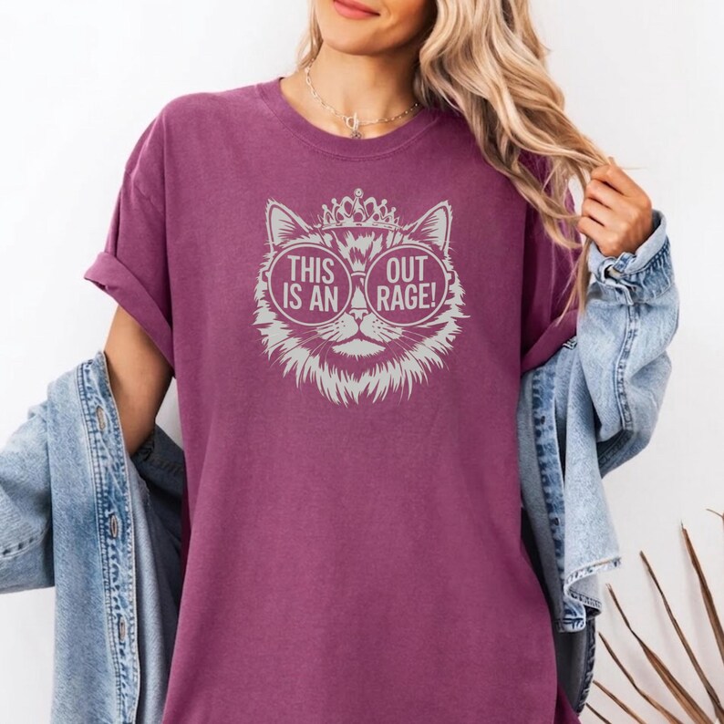 May include: A maroon t-shirt featuring a cat graphic with sunglasses and a crown. The text on the shirt reads "THIS IS AN OUT RAGE!". The design is in a light grey color.