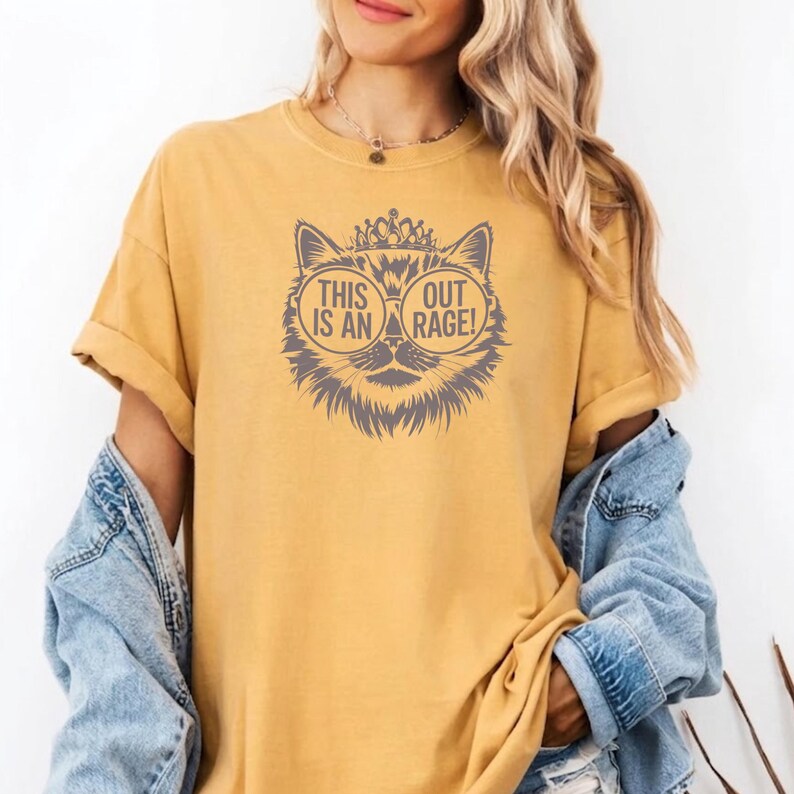 May include: Mustard yellow t-shirt featuring a cat graphic with sunglasses and a crown. The cat graphic has the text "THIS IS AN OUT RAGE!". The shirt has short sleeves and a relaxed fit.