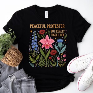 May include: A black t-shirt featuring a colorful botanical illustration of various flowers including a large pink orchid, blue delphiniums, and red poppies. The text reads, "PEACEFUL PROTESTER BUT REALLY PISSED OFF" in a bold, orange-yellow font.