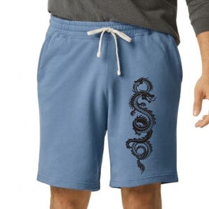 May include: Light blue shorts featuring an elastic waistband with a white drawstring. A black graphic of a coiled dragon is printed vertically on the left leg. The shorts are made of a solid-colored fabric and have a standard knee-length cut.