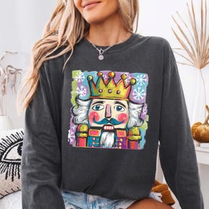 May include: A charcoal gray long-sleeve sweatshirt featuring a vibrant, painterly graphic of a nutcracker wearing a gold crown. The design includes colorful accents, white snowflake patterns, and a stylized artistic background in shades of blue, green, and purple.