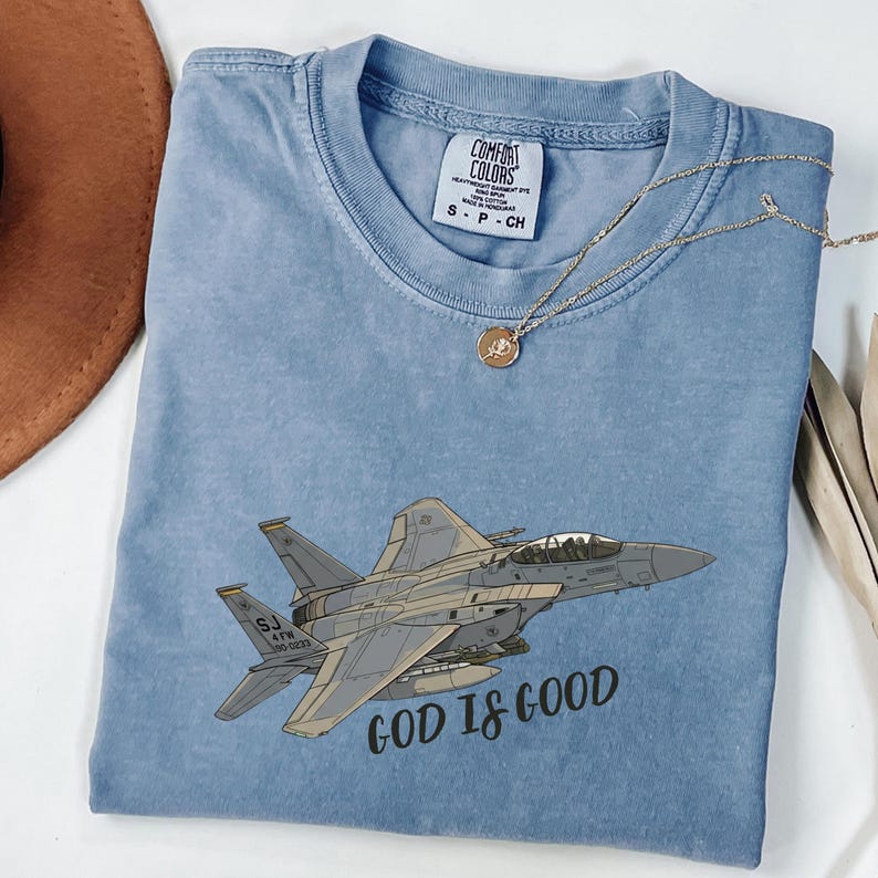 Pu&ograve; includere: T-shirt Comfort Colors azzurra con grafica di un jet da combattimento grigio e la scritta "GOD IS GOOD". La maglietta ha girocollo e maniche corte. Una collana d'oro con un ciondolo &egrave; drappeggiata sulla maglietta.