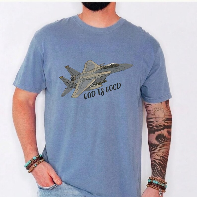Pu&ograve; includere: T-shirt azzurra con un'illustrazione di un jet da combattimento grigio e la scritta "GOD IS GOOD". Il disegno &egrave; stampato su una t-shirt girocollo a maniche corte.